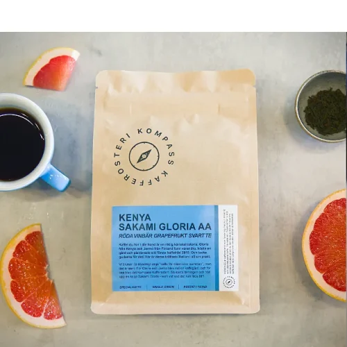En påse Kenya Gloria AA på ett bord, omgiven av grapefrukt, svart te och en kopp kaffe